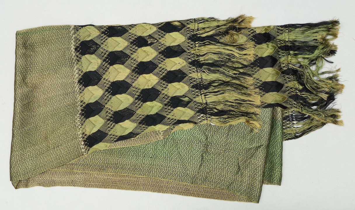 Long, rectangular shawl narrow stripes in green, beige and black. Some of the stripes have ikat-style dots in beige and black. The transverse ends of the shawl are decorated with long fringes with a woven pattern of diamonds in black and green. This is one of the 'Frida Kahlo' garments of Diane Joubert. Antilles, 1950s/1960s (TRC 2023.1219).