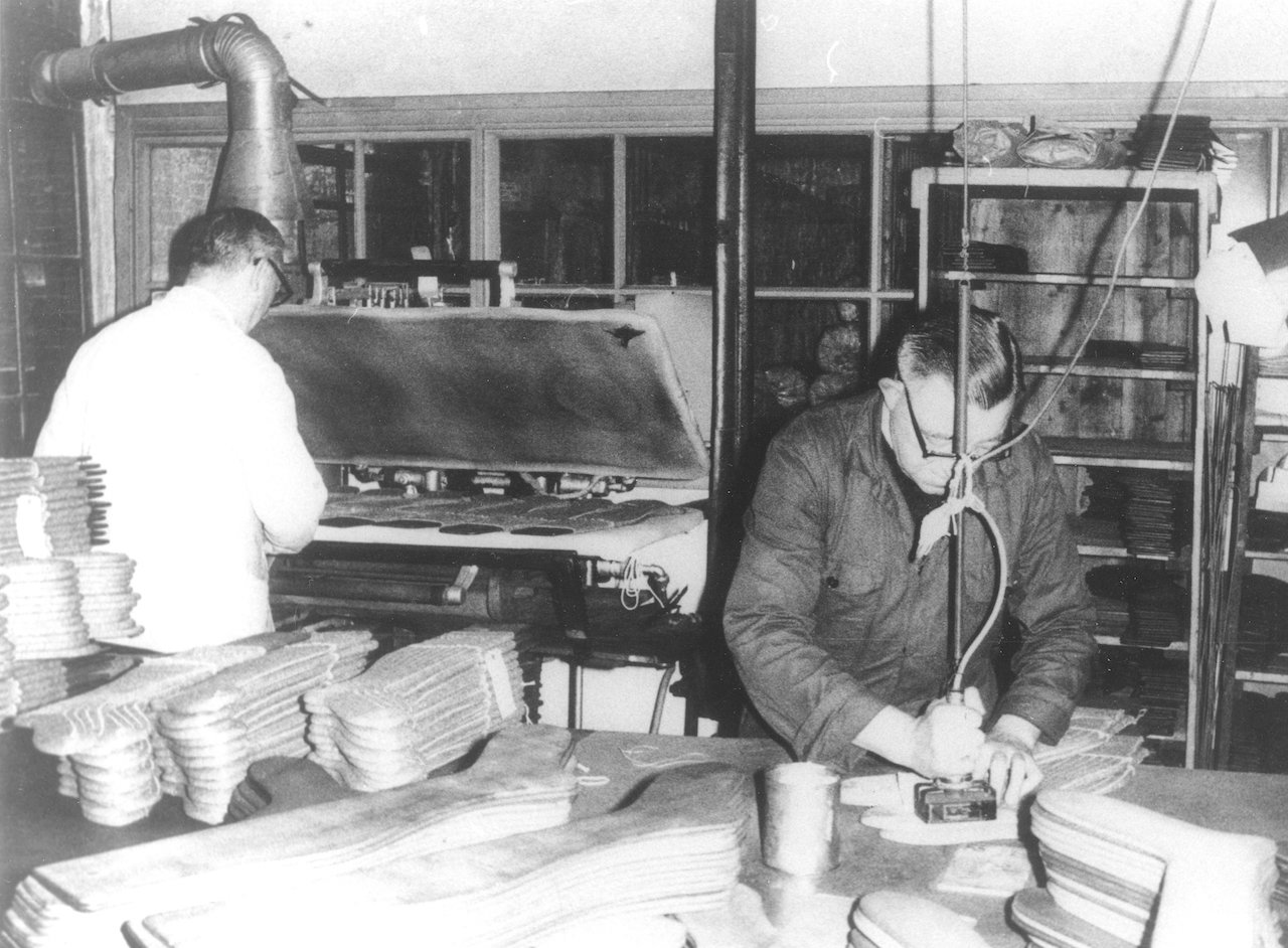 Two men at work in the factory of Van Poot, Leiden, with piles of the wooden forms for making socks and stockings. Date unknown, but the hair style and glasses would indicate a date after 1945.