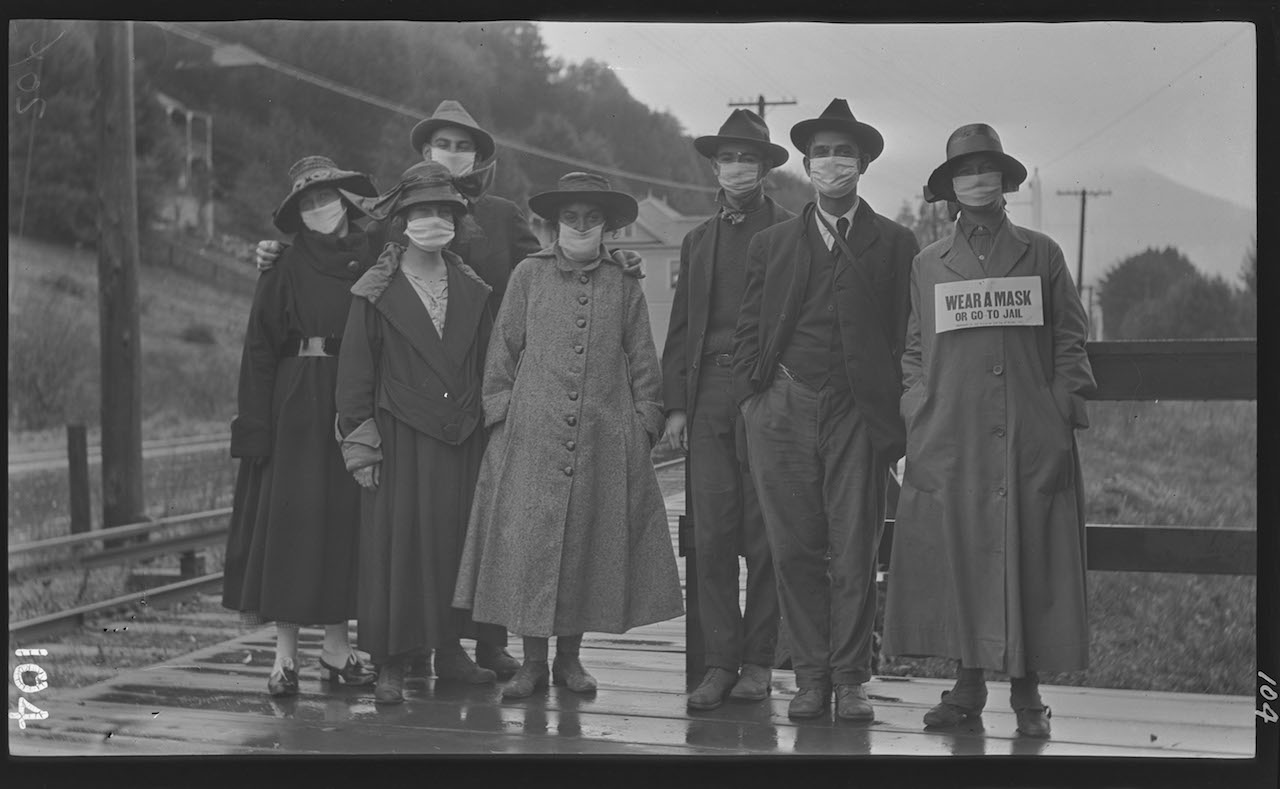A group of people standing outdoors wearing masks over their mouths. This was probably during the Spanish Flu epidemic of 1918. One of the women has a sign in front of her reading 'Wear a mask or go to jail." Courtesy Digital Public Library America.